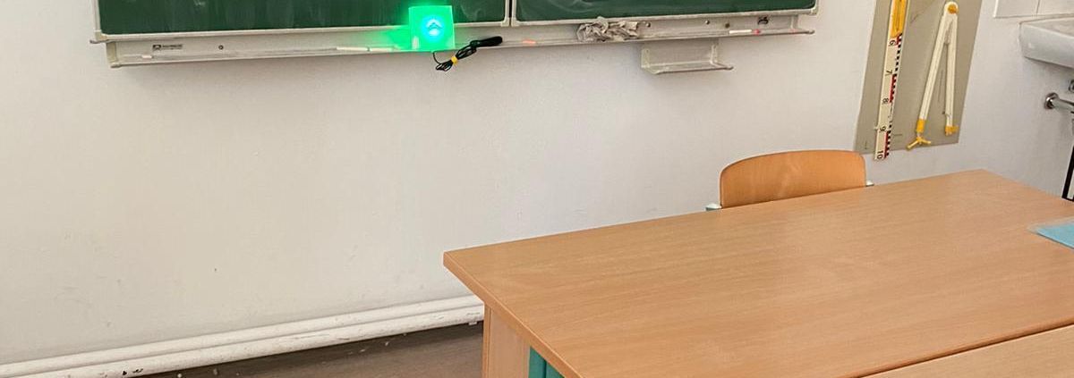 Klassenzimmer mit grüner Tafel, Tischen und Stühlen. Auf der Tafel leuchtet CO2-Ampel grün.