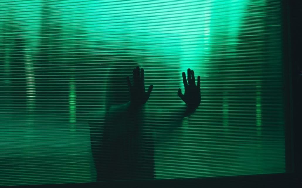 Eine Frau steht hinter einer Wand aus grünem, geriffelten Glas und drückt ihre Hände dagegen. Man kann sie nicht erkennen.